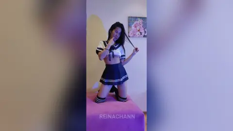 Young woman in a navy sailor cosplay outfit posing playfully in a bedroom setting.
