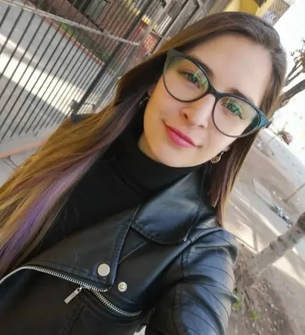 Smiling young woman in a black leather jacket and glasses posing outdoors in natural sunlight.