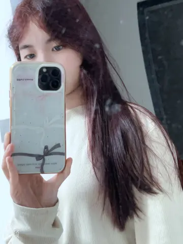 Young woman with long reddish-brown hair taking a mirror selfie in a white ribbed sweater.