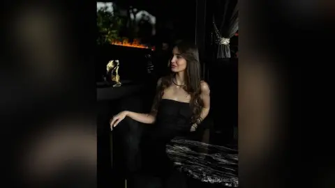 OliviaHardies sits in a dark room wearing a black strapless dress, looking off to the side with her long hair down.