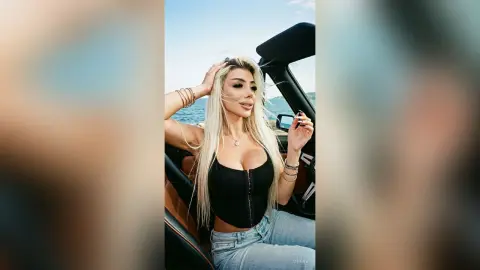 Blonde woman in a black tank top and jeans sitting in a convertible car by the ocean on a sunny day.