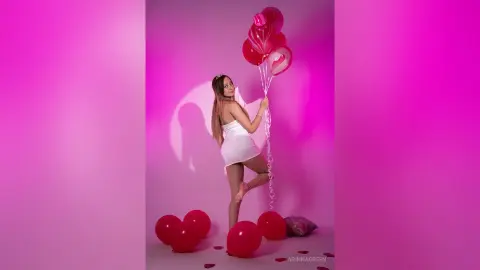 ArinnaGreen poses in a short white dress, holding a bunch of red balloons against a pink background.