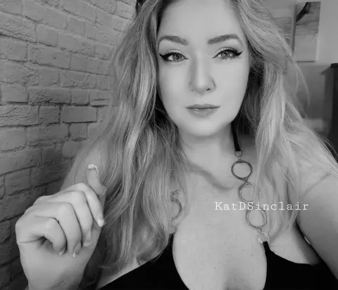 KatDSinclair poses with long blonde hair and winged eyeliner, wearing a black top and a silver ringed choker against a brick wall.