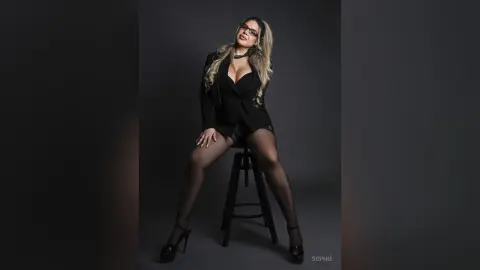 Confident woman in a black blazer and sheer tights posing against a dark studio background.
