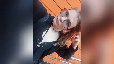 Young woman with glasses in a black outfit posing against an orange wall for a moody portrait.