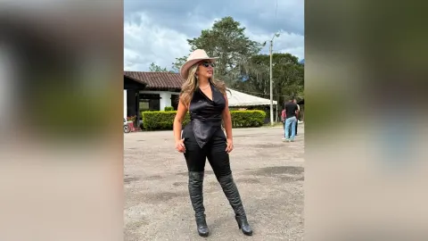 CrisstyBaez — CrisstyBaez stands outdoors in a black halter top, black leggings, and tall black boots while wearing a tan cowboy hat.