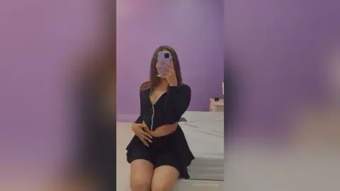 Young woman in a black long-sleeve crop top and skirt posing for a mirror selfie in a purple room.