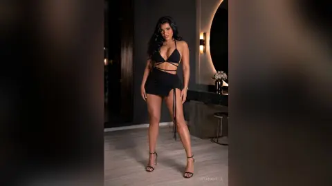 DayanaHille stands in a black lace teddy, showing off her long legs and dark hair in a dimly lit room.