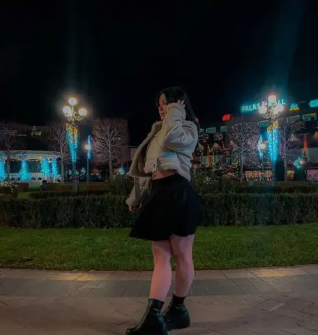 Young woman in a beige jacket and black skirt standing in a city park at night near glowing mall lights.