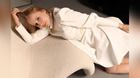 Elegant woman in a white long-sleeve dress and gold jewelry reclining on a plush cream rug in a bright room.