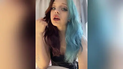 Young woman with split-dye burgundy and blue hair wearing a black top in a soft, moody indoor setting.