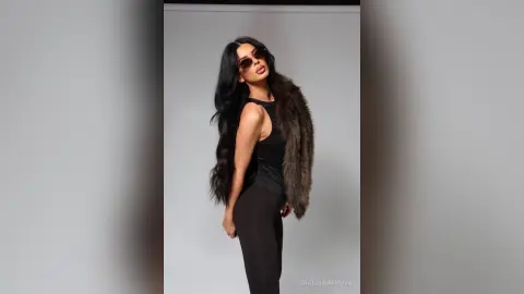Stylish woman in a black tank top and brown faux fur coat posing confidently against a grey studio background.