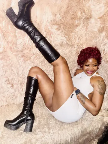 Smiling woman in white bodysuit and black platform boots lounging on a fluffy pink faux fur rug.