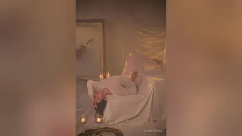 AlanaPearce lies back on a white sheet in a dimly lit room, wearing a white long-sleeve top and white pants, surrounded by soft candlelight.