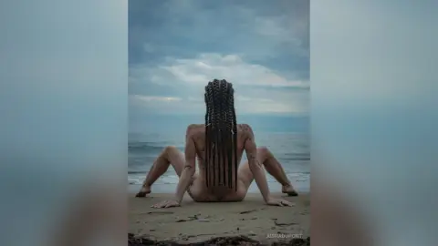 Nude woman with long braids sitting on a sandy beach under a moody, overcast sky facing the ocean.