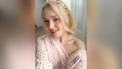 Smiling blonde woman in a cozy pink knit sweater wearing a pearl headband in a bright indoor setting.