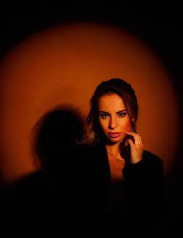 Moody portrait of a blonde woman in a black blazer against a warm orange spotlight background.