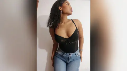Confident woman in a black sheer bodysuit and blue jeans posing against a white wall.