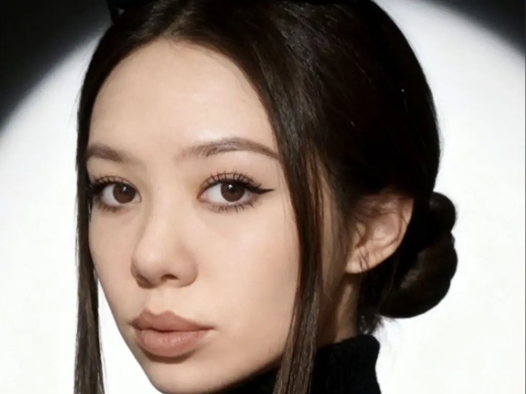 Close-up portrait of a woman with dark hair and winged eyeliner looking into the camera against a bright background.