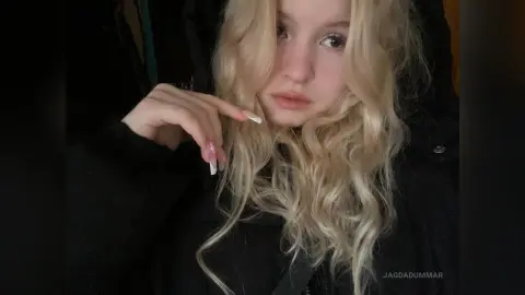 Blonde woman in a black hoodie with long wavy hair posing with a moody expression indoors.