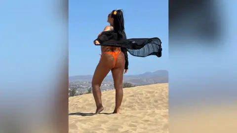 MissyJolie stands on a sand dune in an orange bikini, looking over her shoulder with a black mesh cover-up blowing in the wind.