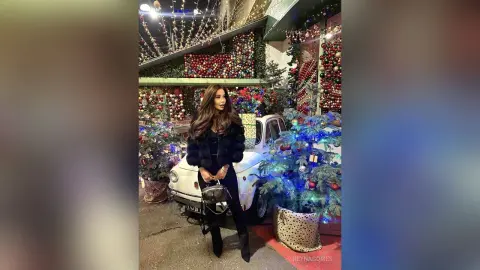 ReynaGomes stands outside wearing a black furry jacket and black pants, posing next to a white car and decorated Christmas trees.