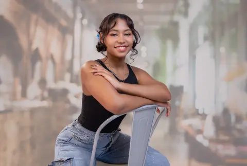 Smiling young woman in a black tank top and jeans sitting in a bright, modern indoor setting.