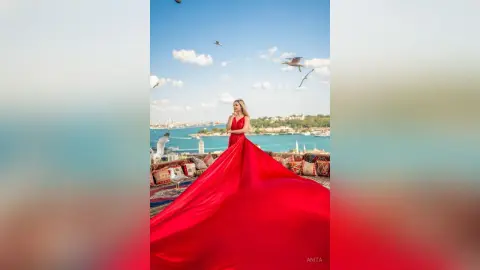 Anita wears a long red dress with a flowing train while standing outdoors overlooking a blue sea and coastline.