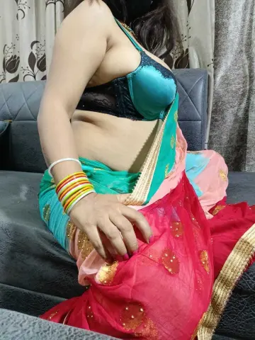 Woman in a colorful pink and teal traditional saree sitting on a grey sofa in a cozy indoor setting.
