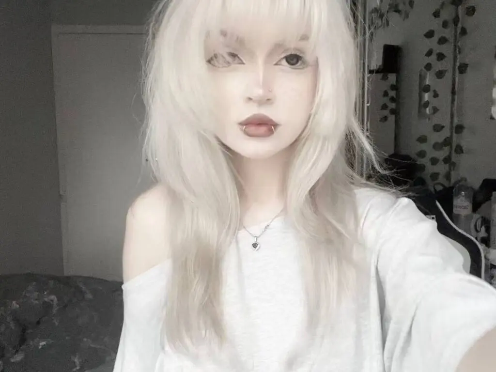 LeticiaFinello poses with long blonde hair and a lip piercing, wearing a white off-the-shoulder shirt in a dimly lit room.