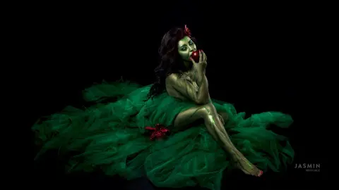 MissyJolie poses in a voluminous green tulle gown, her skin or body painted green, while holding a red apple to her lips.
