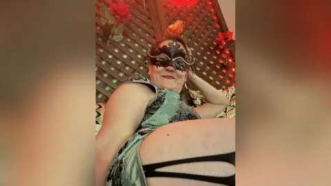 Smiling woman in a silver sequin dress and ornate masquerade mask relaxing in a room with red roses.