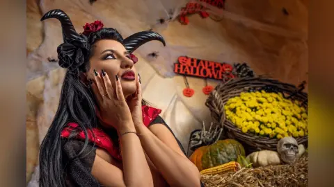 MissyJolie poses with her hands against her face, wearing black hair accessories and a red and black outfit against a Halloween-themed background.