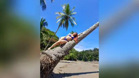 Redheaded woman in a blue bikini relaxing on a fallen palm tree trunk at a sunny tropical beach.