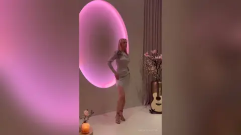 KeilaTouchton poses in a tight grey dress standing next to a large glowing pink ring light.