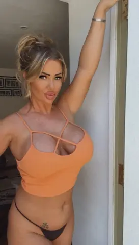 Curvy blonde woman in an orange crop top and bikini posing confidently in a bright doorway.