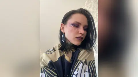 AndreaHazel looks toward the side with dark hair and purple lipstick, wearing a black and gold patterned shirt.