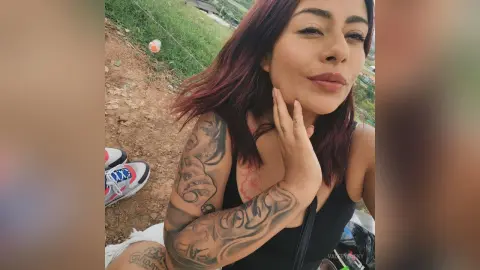 Smiling woman with red hair and arm tattoos wearing a black tank top in a sunny outdoor field setting.
