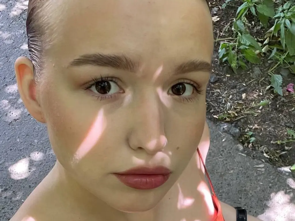 Close-up portrait of a young woman with natural makeup and slicked-back hair in a sunny outdoor garden setting.
