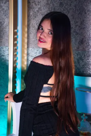 Smiling young woman with long brown hair in a black backless dress posing in a moody, teal-lit room.