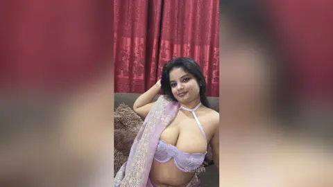 Young woman in a lavender lace lingerie set and sheer wrap posing indoors against red curtains.