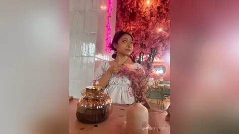 Young woman in a white blouse sitting at a cafe table near pink cherry blossom decor in a dreamy atmosphere.