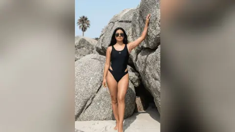 Stylish woman in a black one-piece swimsuit posing against large rocks on a sunny beach.
