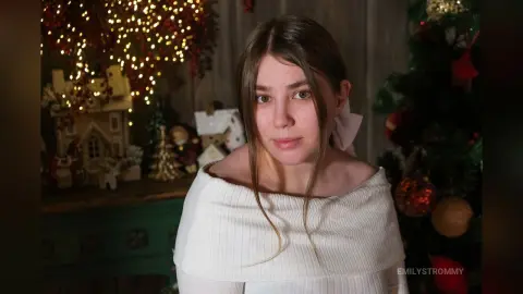 Young woman in a white off-the-shoulder top smiling in a cozy room with festive Christmas lights and decor.