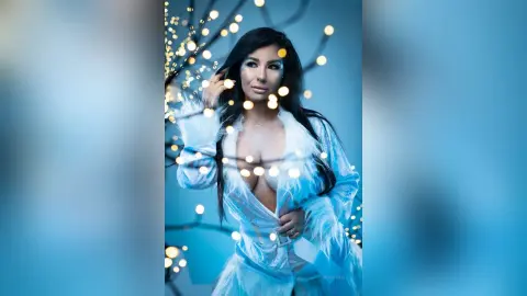 MissyJolie poses in a light blue feathered robe, running her hands through her long dark hair against a blue background with twinkling lights.