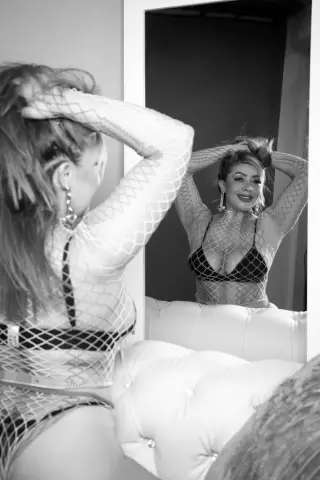 Glamorous woman in a white fishnet bodysuit posing in a mirror for a stylish black and white portrait.