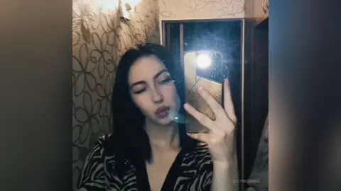 Young woman with long dark hair in a patterned black top taking a moody mirror selfie in a dimly lit room.