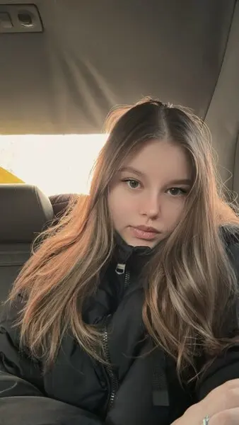 Enchaantress poses for a selfie, wearing a maroon top with her long brown hair styled straight down against a neutral wall.