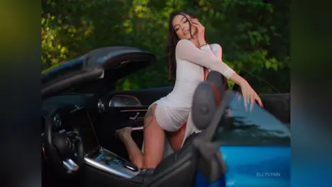 Elegant woman in a white dress posing in a blue convertible car against a lush green garden background.