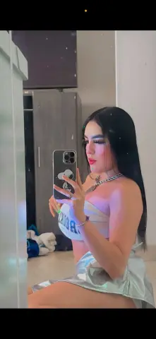 Young woman with long dark hair in a white crop top and silver skirt taking a mirror selfie in a modern room.